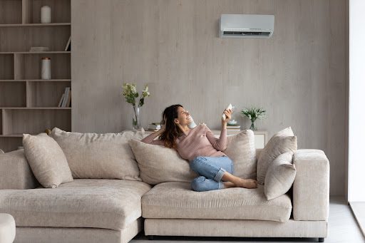 A happy woman relaxing on her big couch as she turns on her home’s air conditioning system using a remote.